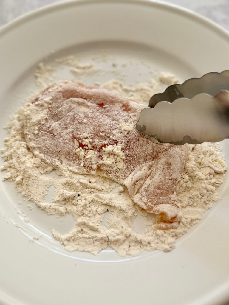 chicken breast coated in flour with tongs on white plate
